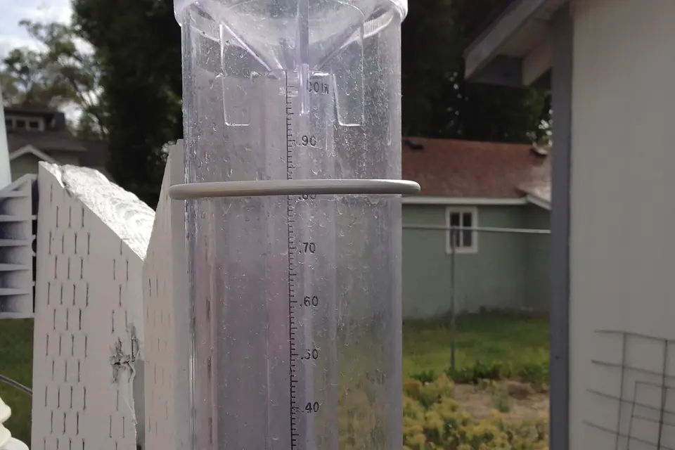 Un pluviomètre manuel en plastique transparent avec son tube intérieur gradué, fixé à une clôture de jardin
